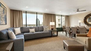 A modern hotel room with a large gray sofa, a small table, a chair, and a large window offering a city view. - Doubletree by Hilton McLean Tysons (McLean)