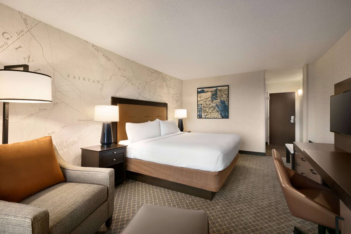A hotel room with a large bed, a desk, a chair, a lamp, and a map on the wall.