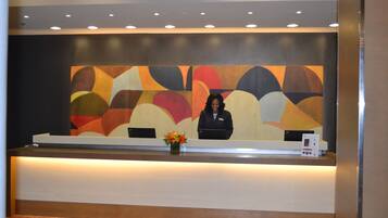 A reception desk with a receptionist, a colorful abstract painting on the wall, and a vase with flowers.