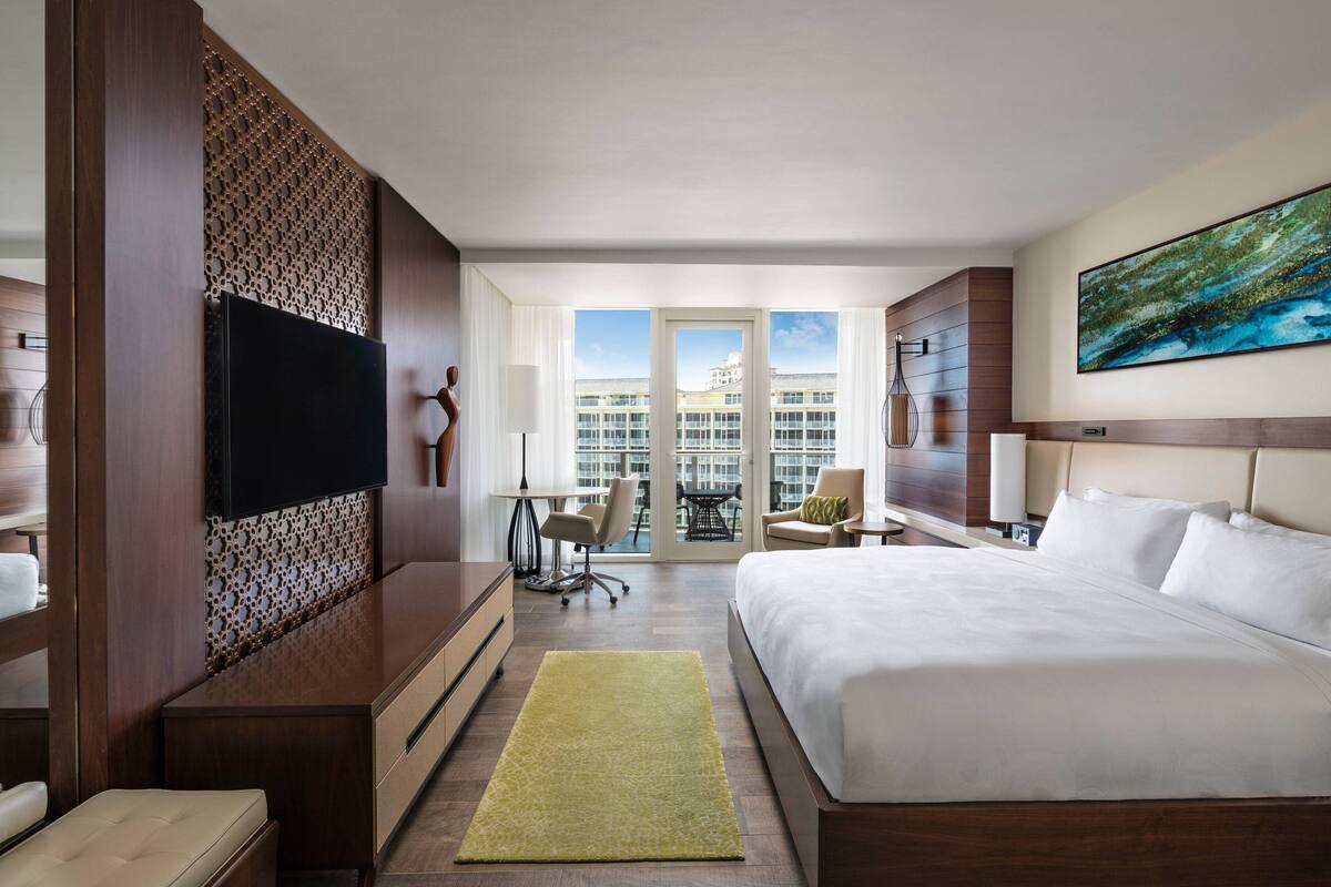 A hotel room with a large bed, a TV mounted on a wooden panel, a balcony with a view of buildings, and a painting on the wall.