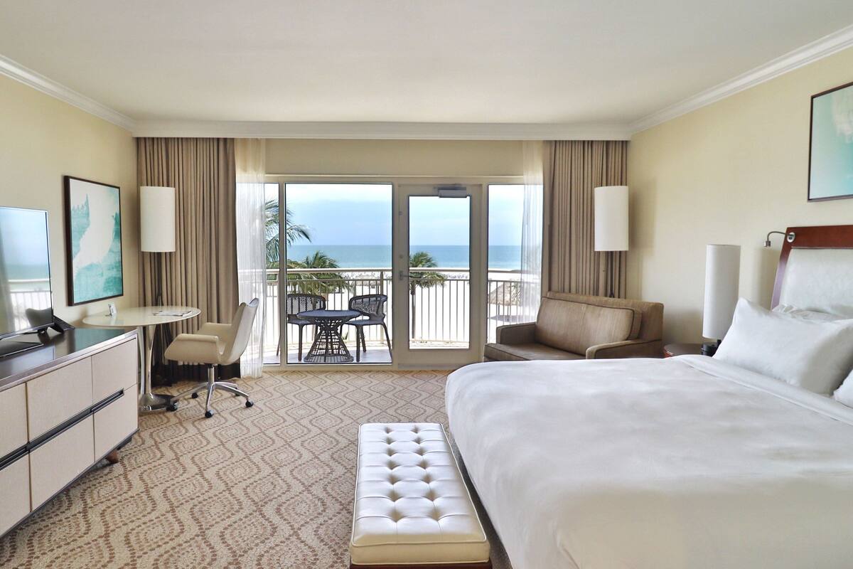 A hotel room with a large bed, a desk, a chair, a small table, and a balcony with a view of the ocean.