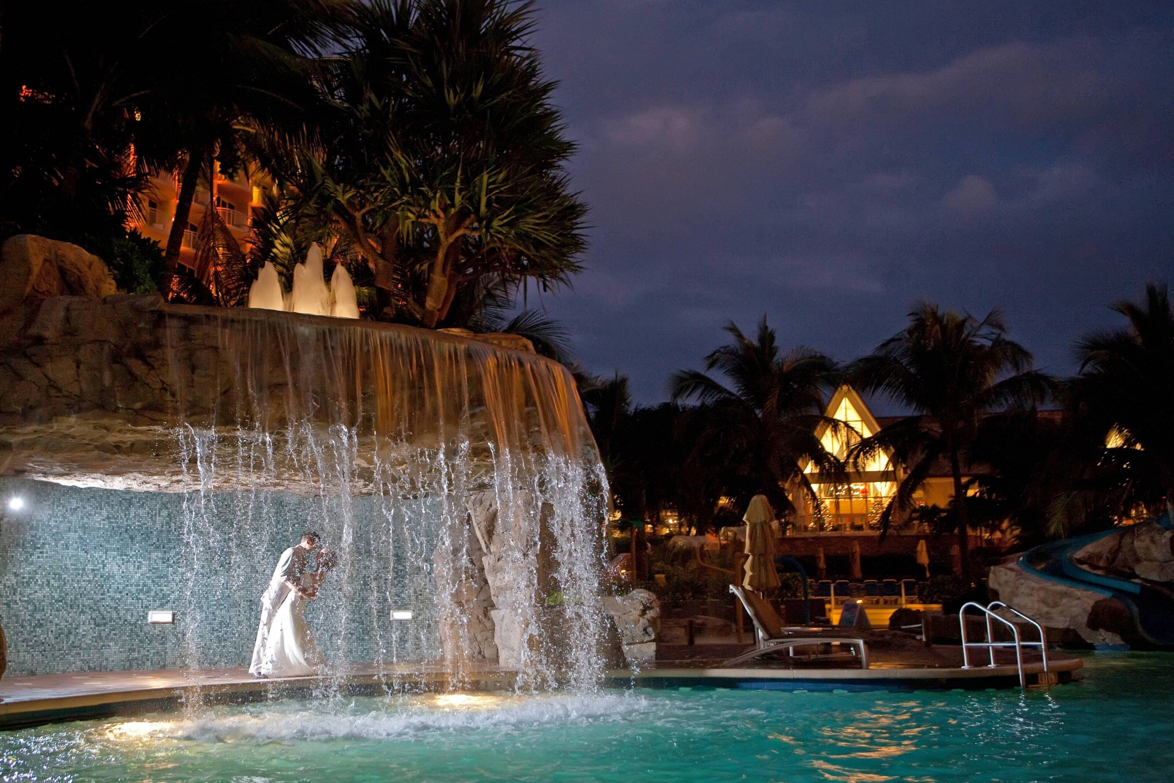 Scena notturna in un resort con cascata, piscina e un edificio con facciata illuminata.
