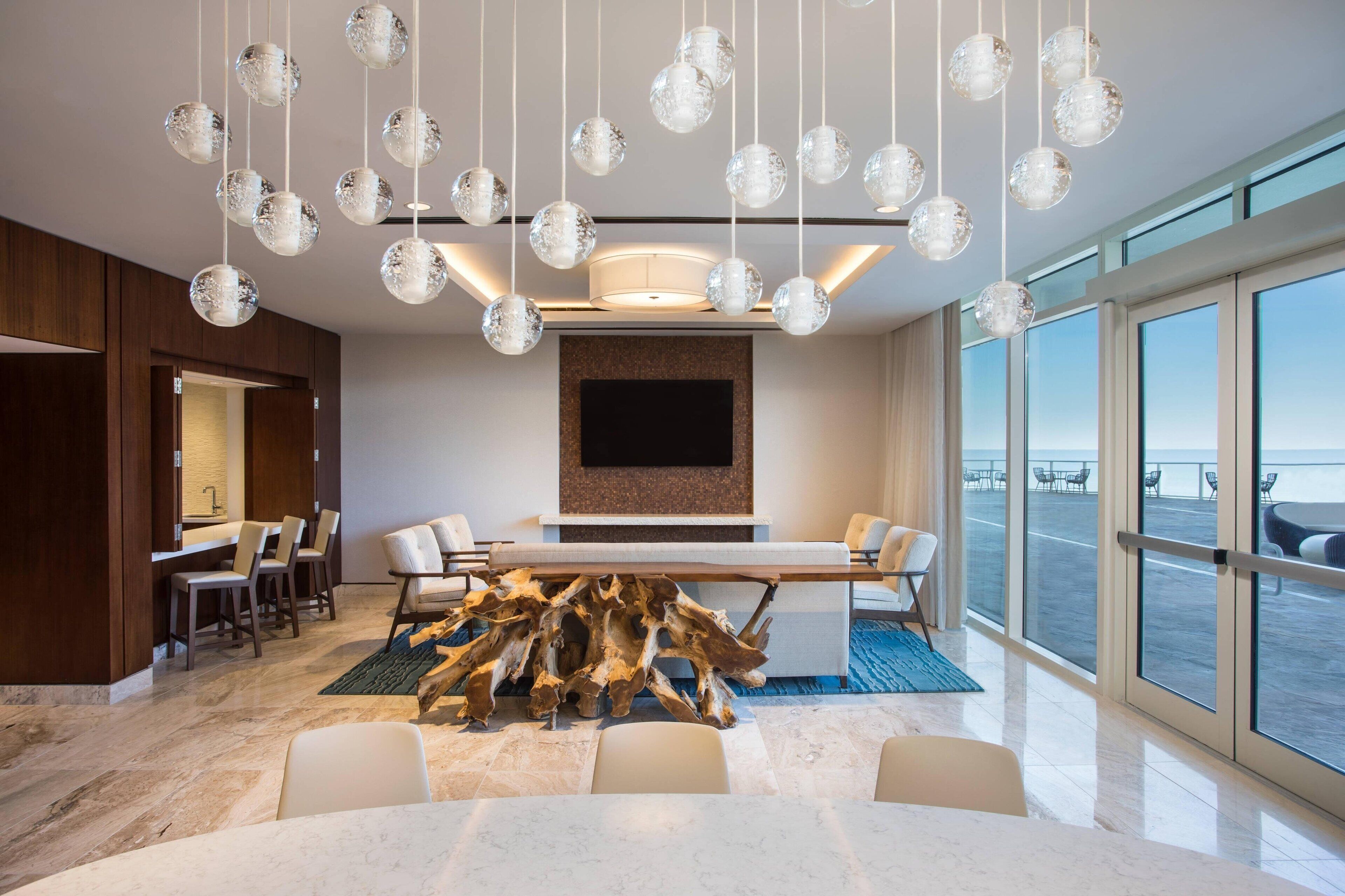 Une chambre d’hôtel moderne dotée d’une grande table à manger, de sièges confortables, d’un téléviseur fixé au mur et offrant une vue sur la mer grâce à de grandes fenêtres.