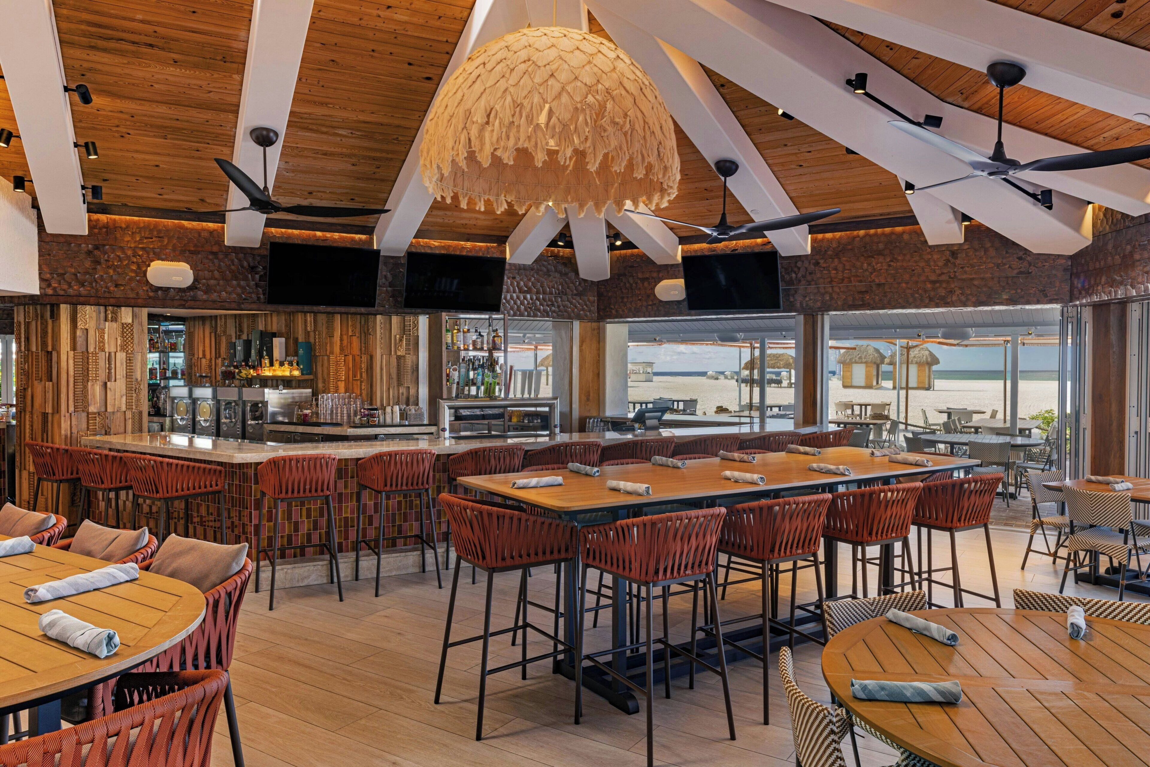 Een modern restaurant met houten balken in het plafond, een centraal bargedeelte en diverse zithoeken, waaronder ronde tafels en hoge stoelen.