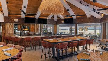 A modern restaurant with wooden ceiling beams, a central bar area, and multiple seating options including round tables and high chairs.