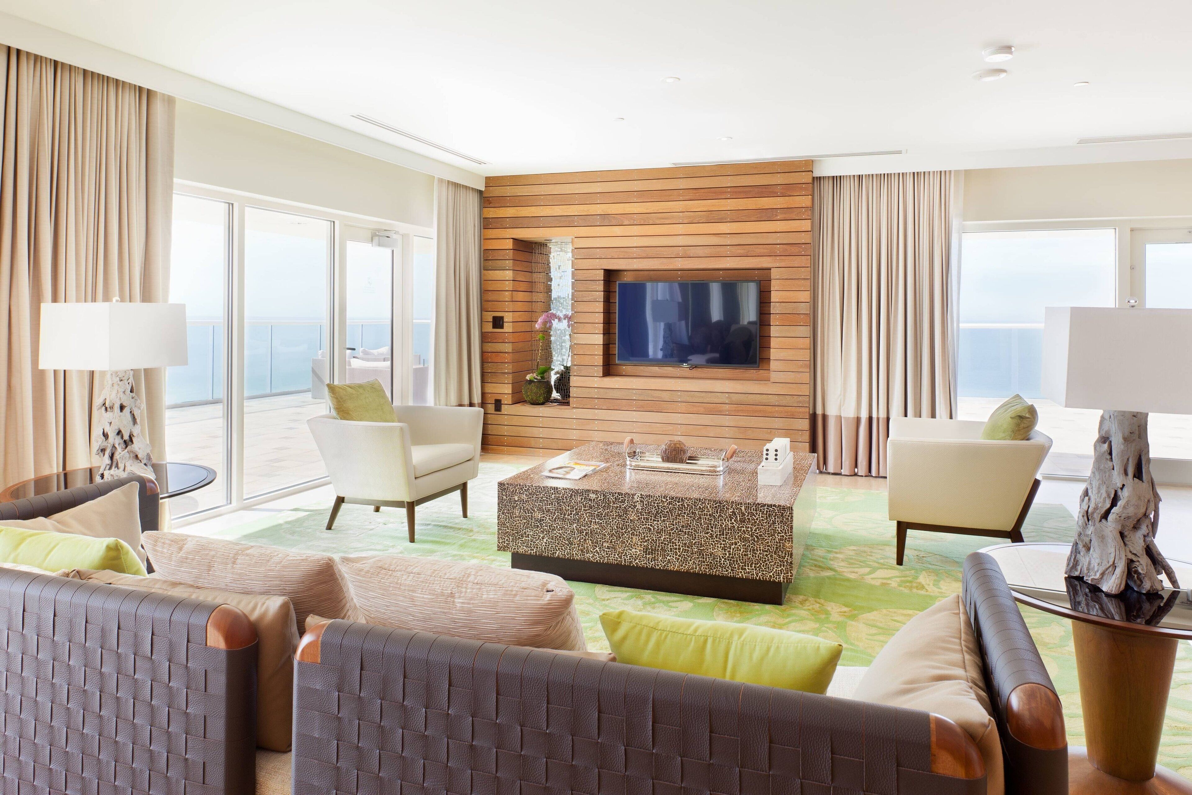 Une chambre d’hôtel moderne dotée d’un grand téléviseur fixé sur un mur en bois, d’un coin salon avec deux fauteuils et un canapé, et d’un balcon avec vue.