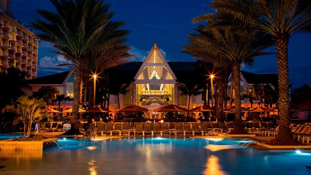 Un hotel con piscina, palmeras y un edificio iluminado al anochecer.