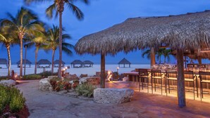 Un área de bar en la playa con sombrillas de paja, palmeras y vista al océano al atardecer.