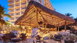Un salón junto a la playa con mobiliario de mimbre, techo de paja y vista a un hotel de gran altura al fondo.