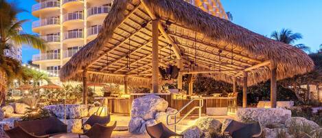 A beachside lounge with wicker furniture, a thatched roof, and a view of a high-rise hotel in the background.