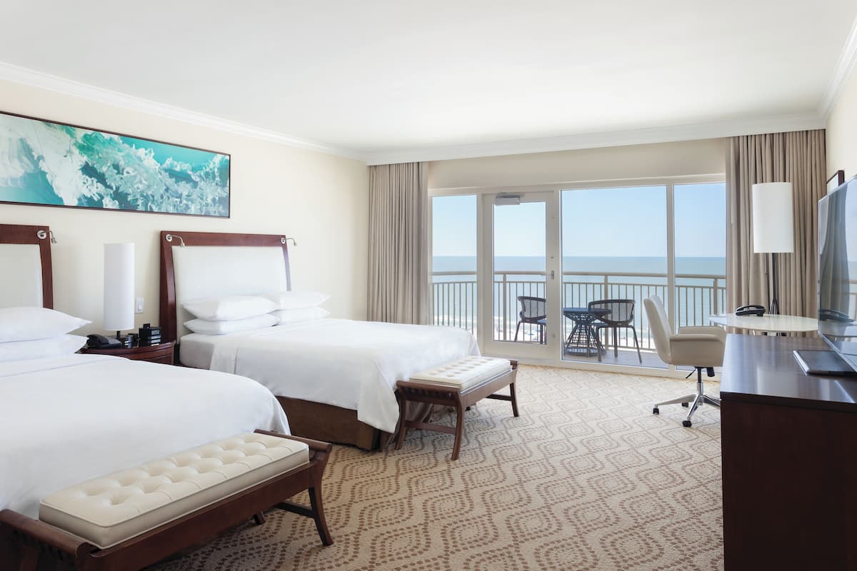 A hotel room with two beds, a desk, a chair, a TV, and a balcony with a view of the sea.