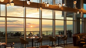 Un restaurante con mesas dispuestas para comer, amplias ventanas que ofrecen vistas al atardecer sobre el océano y lámparas colgantes.