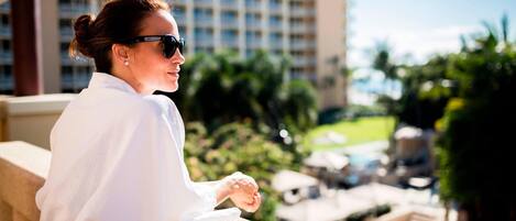 A person leaning on a balcony railing overlooking a pool area with lounge chairs and a building in the background.