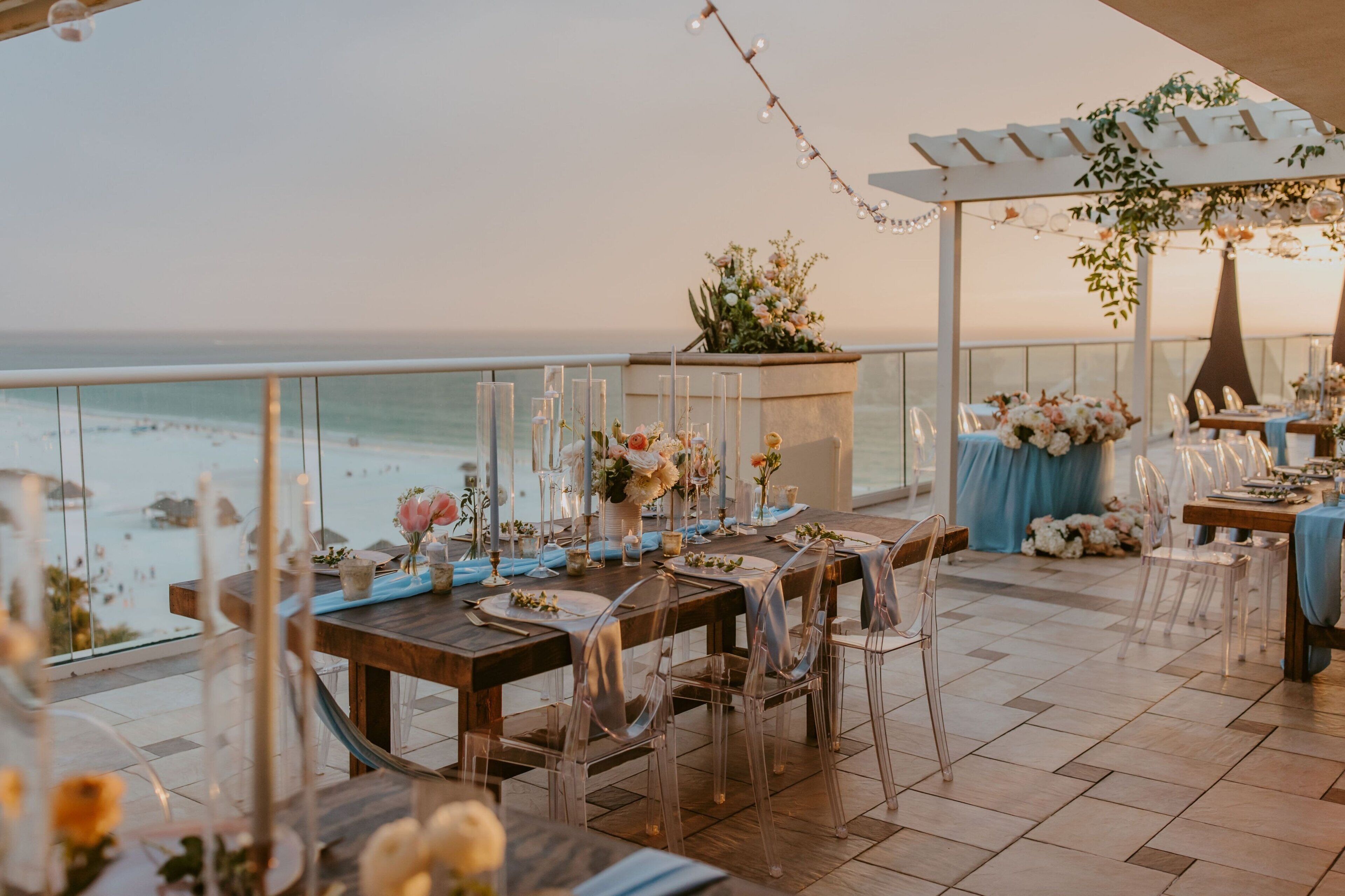 Un espace de restauration en plein air offrant une vue sur l’océan, avec des chaises en verre transparent, des tables en bois et des compositions florales en guise de décoration.
