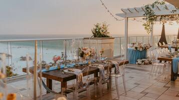 Un espace de restauration en plein air offrant une vue sur l’océan, avec des chaises en verre transparent, des tables en bois et des compositions florales en guise de décoration.