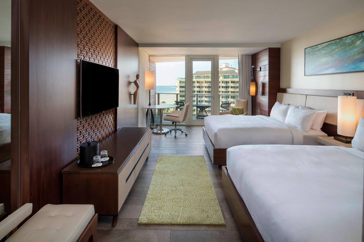 A hotel room with two beds, a TV, a desk with a lamp, a chair, and a view of the ocean.
