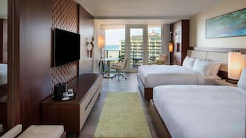 A hotel room with two beds, a TV, a desk with a lamp, a chair, and a view of the ocean.