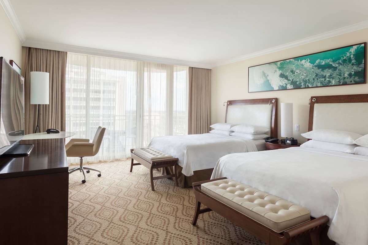 A hotel room with two beds, a desk, a chair, a bench, and a large window with sheer curtains.