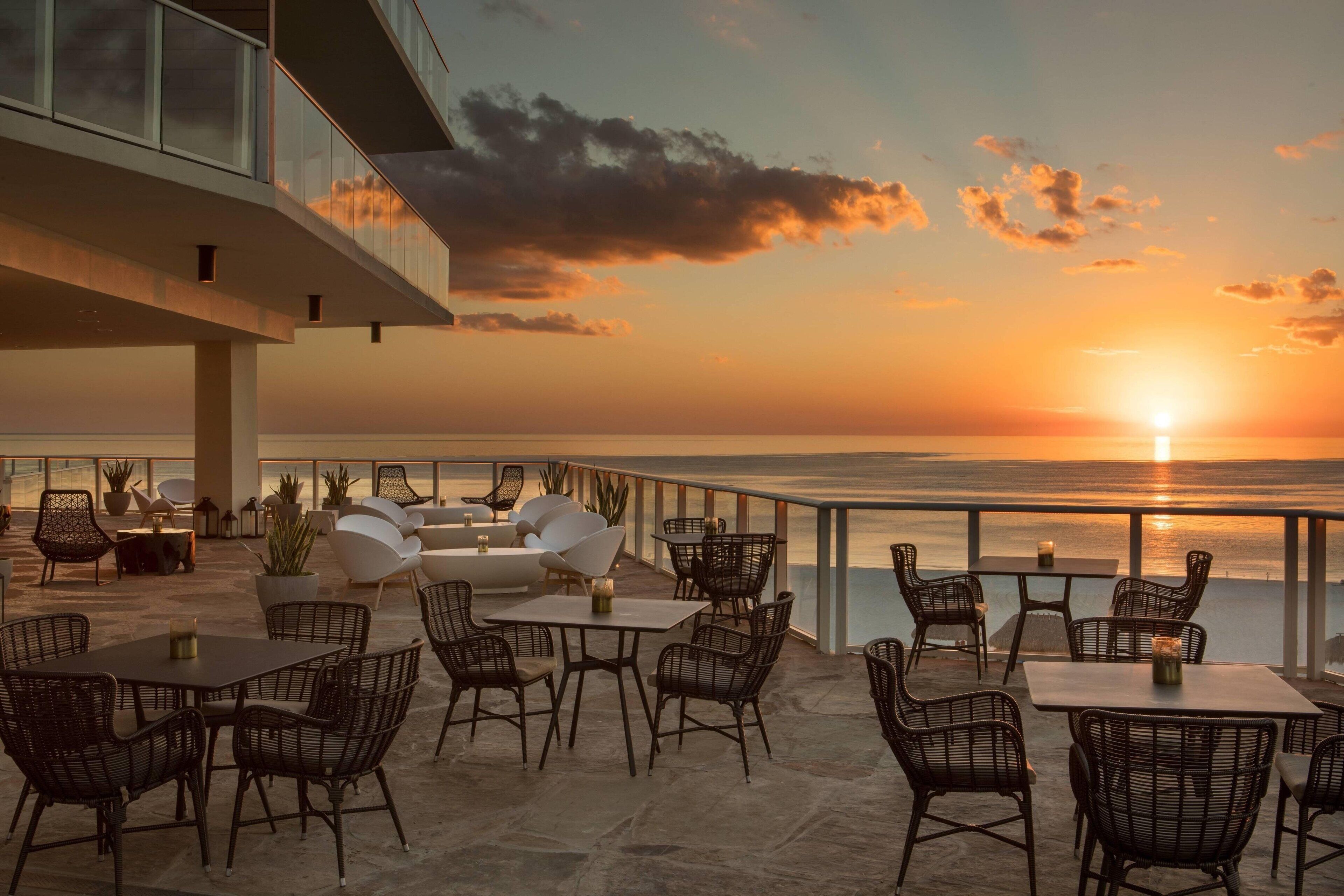 An outdoor seating area with tables and chairs, overlooking a sunset over the water.