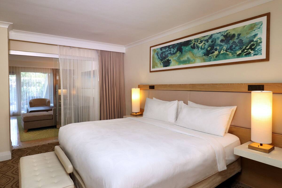 A hotel room with a large bed, two bedside lamps, a headboard, a painting on the wall, a chair, and a view of another room through a glass door.