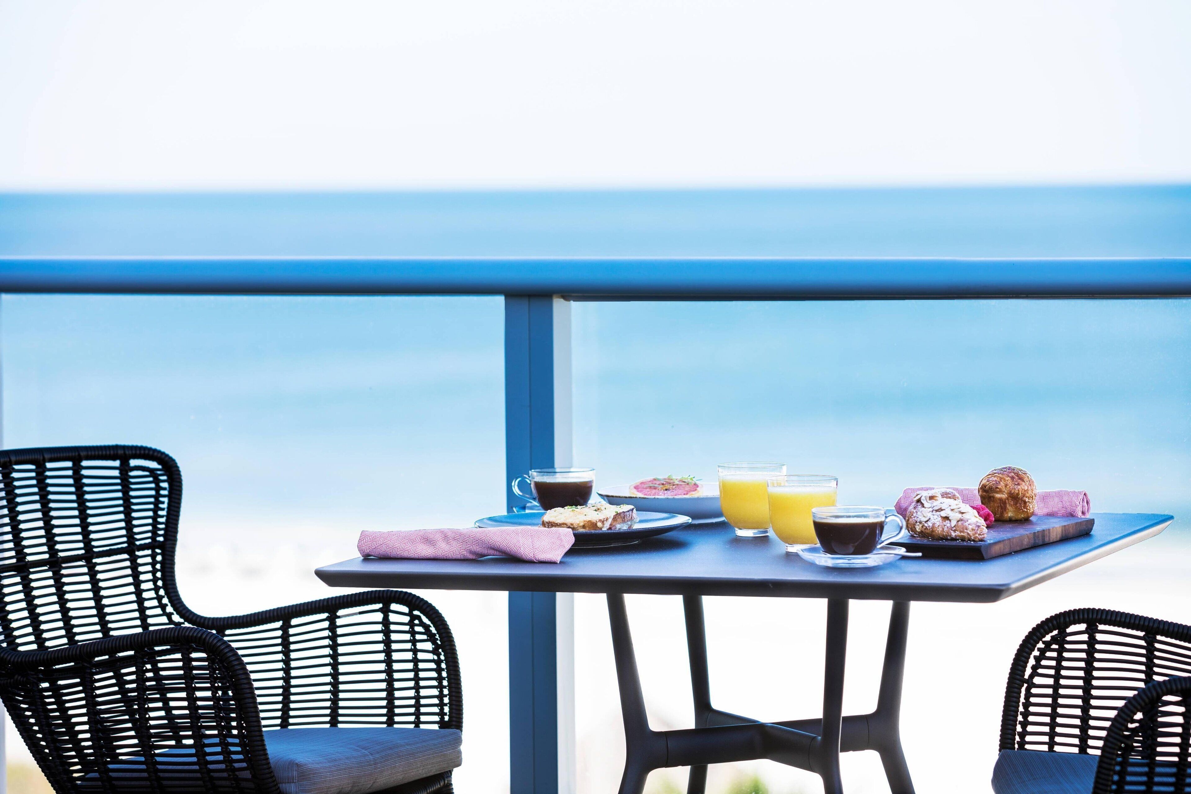 Un balcon avec une table préparée pour le petit-déjeuner, comprenant des viennoiseries, du café et du jus de fruits, donnant sur une étendue d’eau.