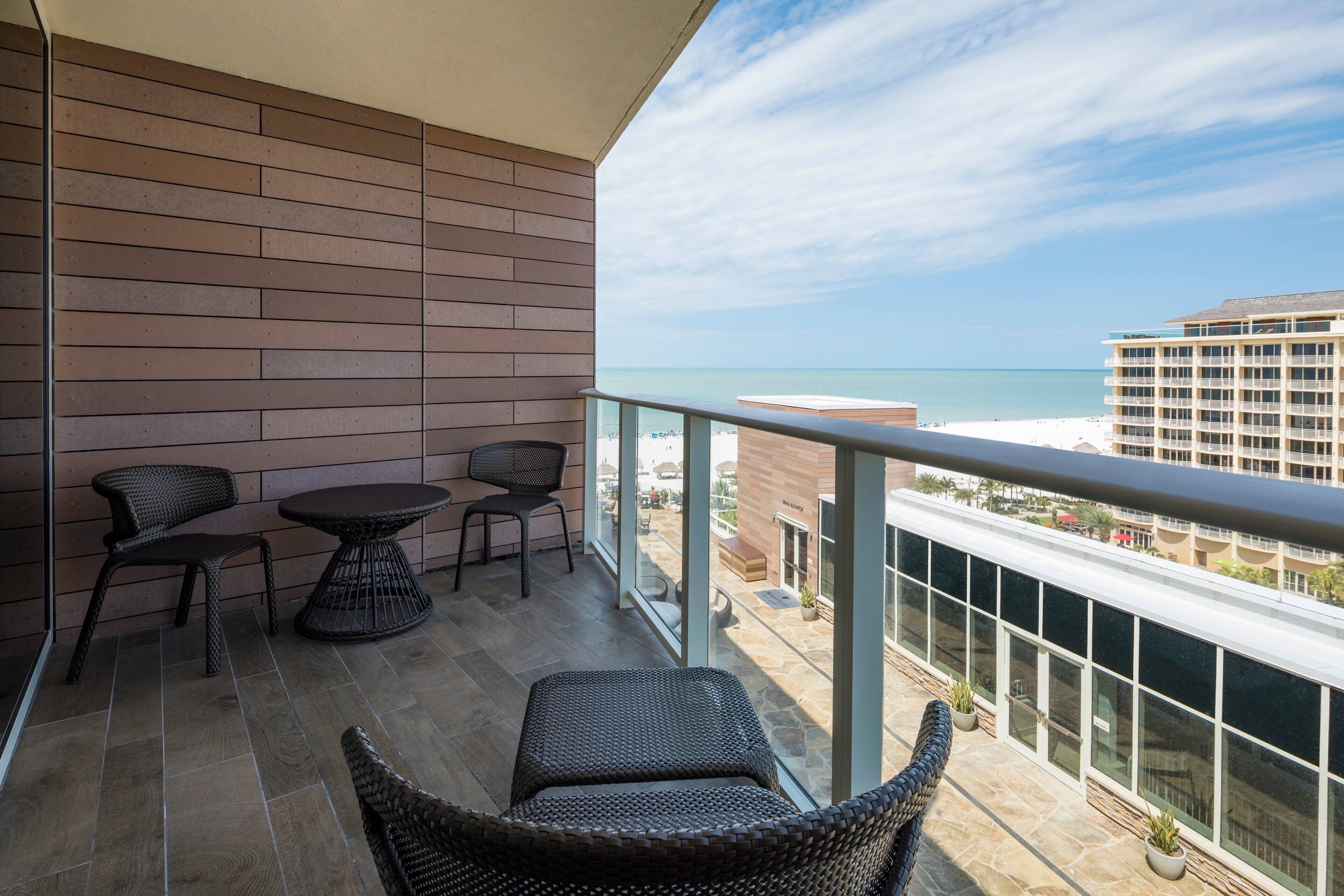 A balcony with wicker furniture, a glass railing, and a view of the ocean and buildings.