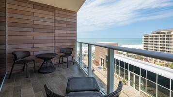 A balcony with wicker furniture, a glass railing, and a view of the ocean and buildings.