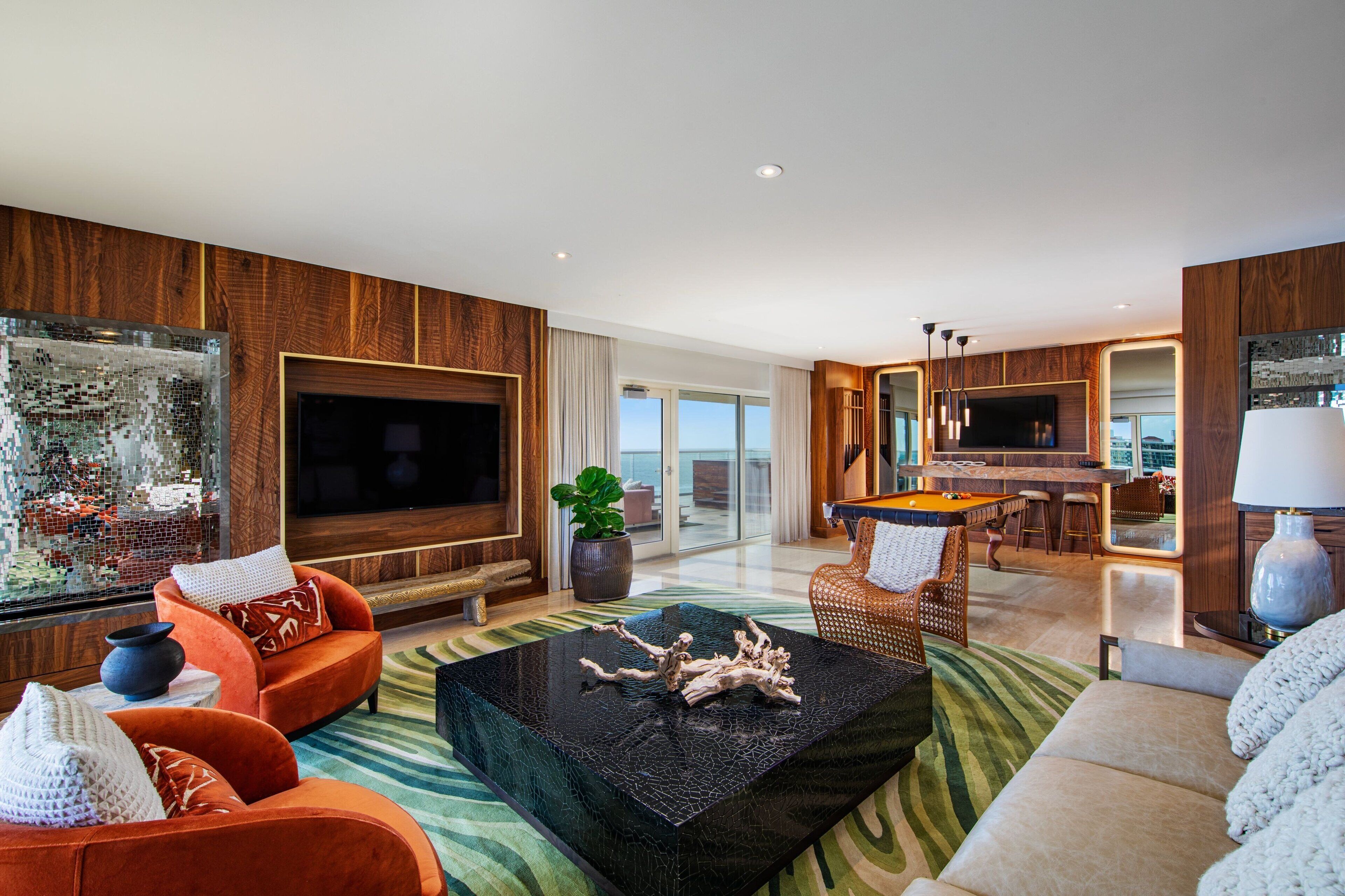 A modern living room with a large flat-screen TV, orange sofas, a coffee table, and a pool table in the background.