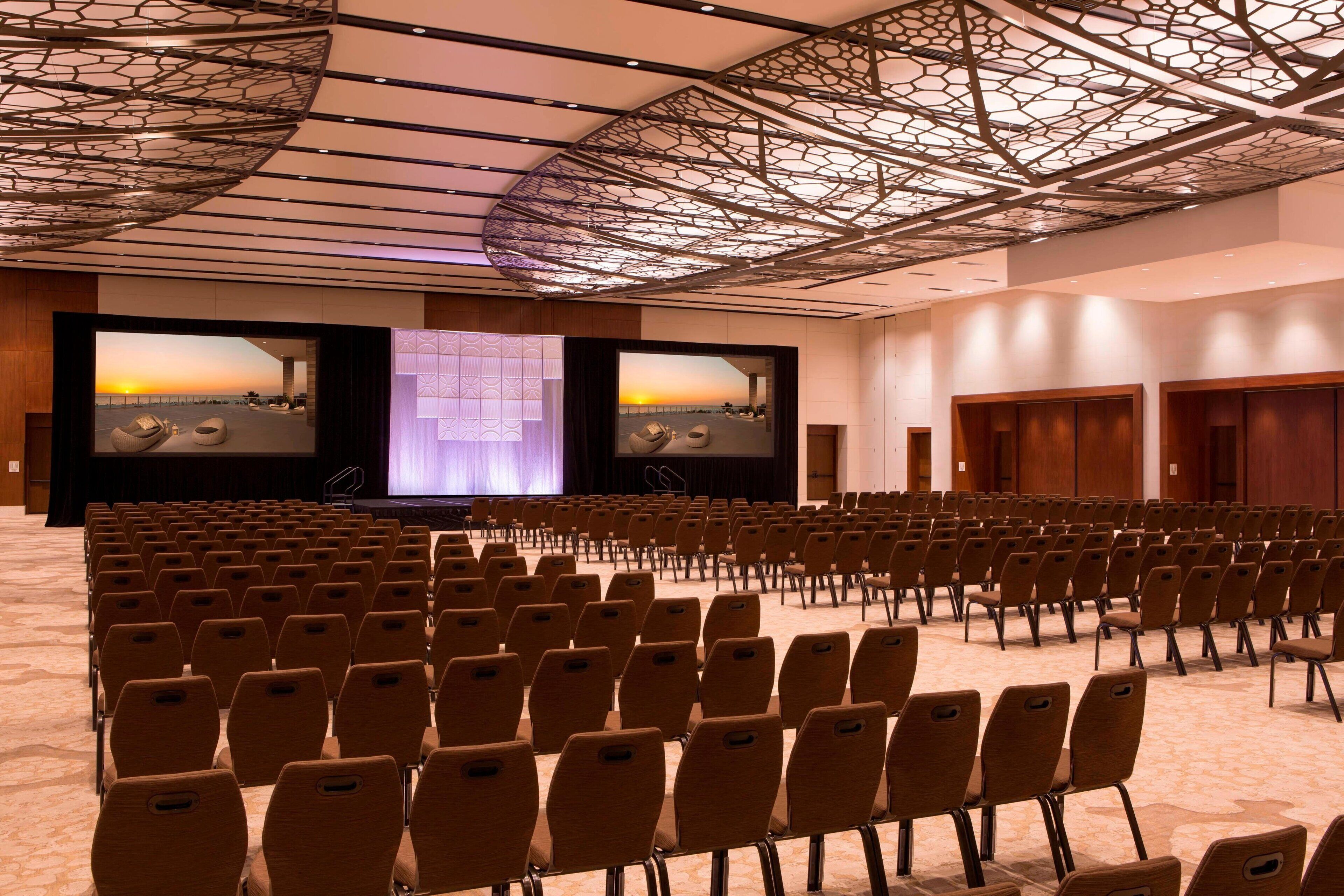 Une salle de conférence avec des rangées de chaises, un grand écran et un plafond orné d’un motif géométrique.