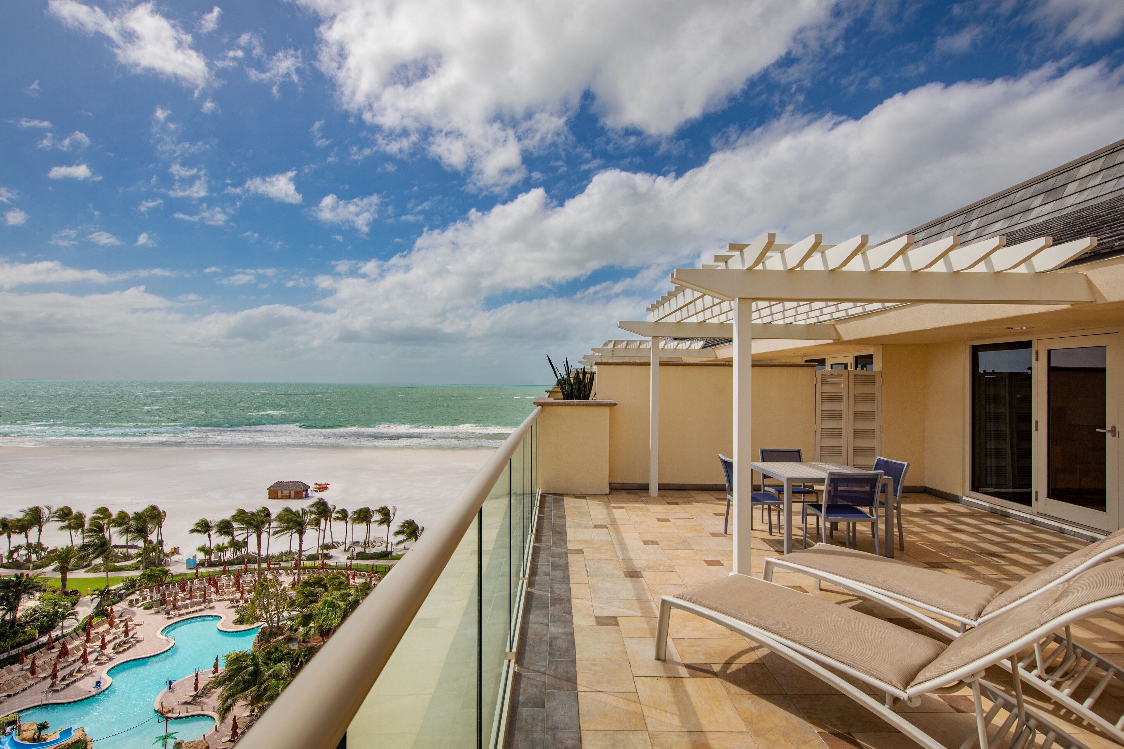 A balcony with a view of the beach and pool, a table with chairs, and a covered area with a pergola.