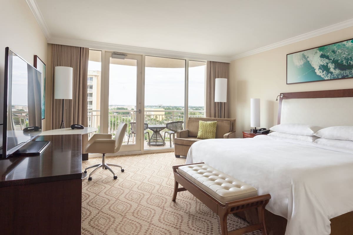 A hotel room with a large bed, a desk with a computer, a chair, a small table, and a balcony with a view.