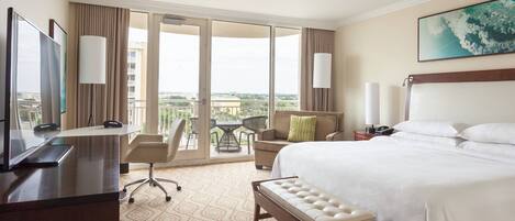 A hotel room with a large bed, a desk with a computer, a chair, a small table, and a balcony with a view.