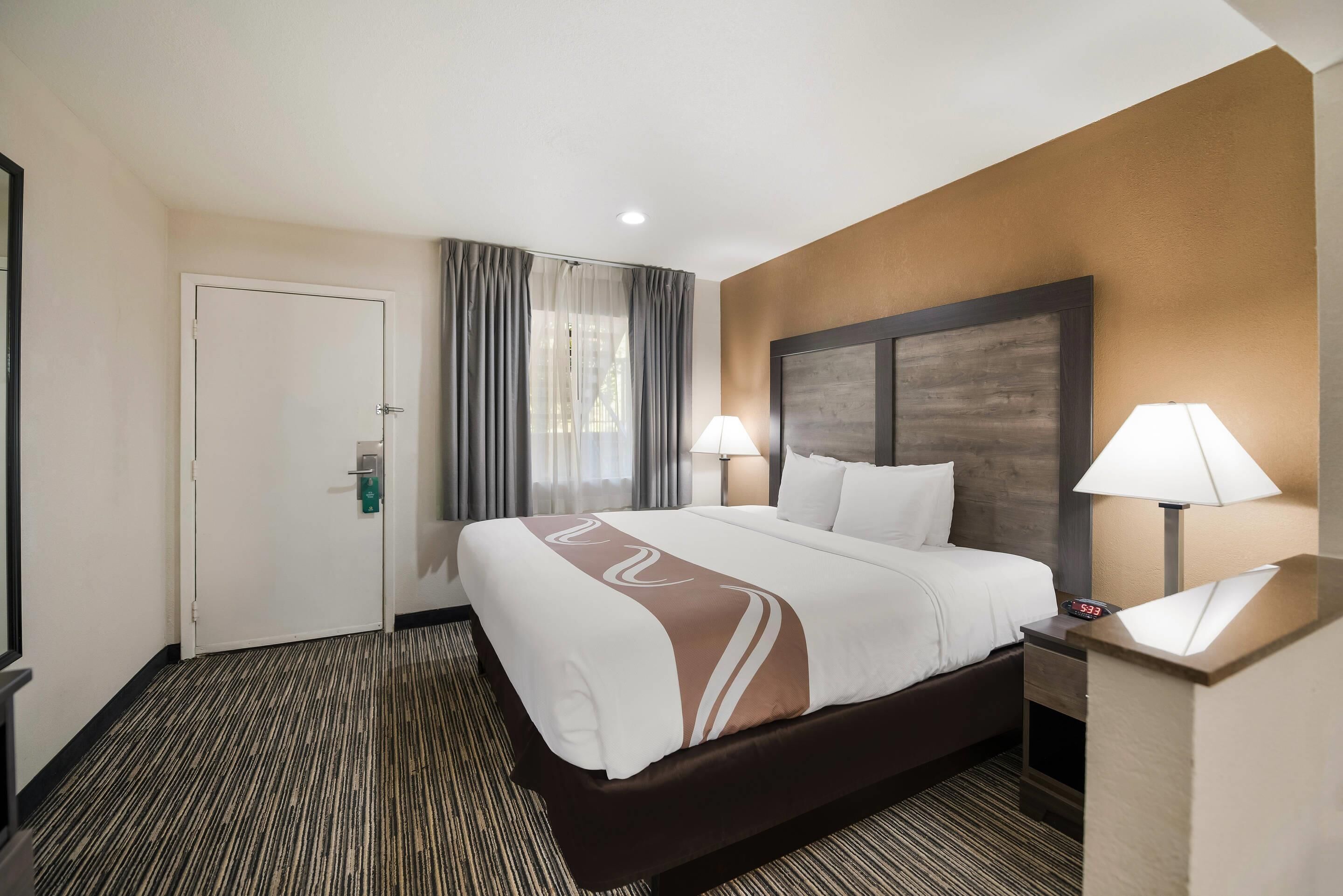 A hotel room with a large bed, two bedside lamps, a nightstand, a door, and a window with curtains.