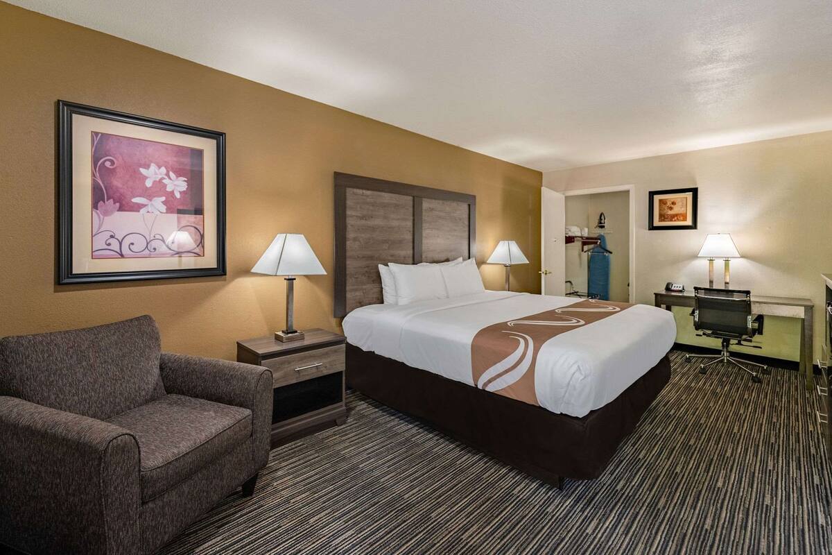 A hotel room with a large bed, two bedside tables with lamps, a chair, a desk, and a framed picture on the wall.