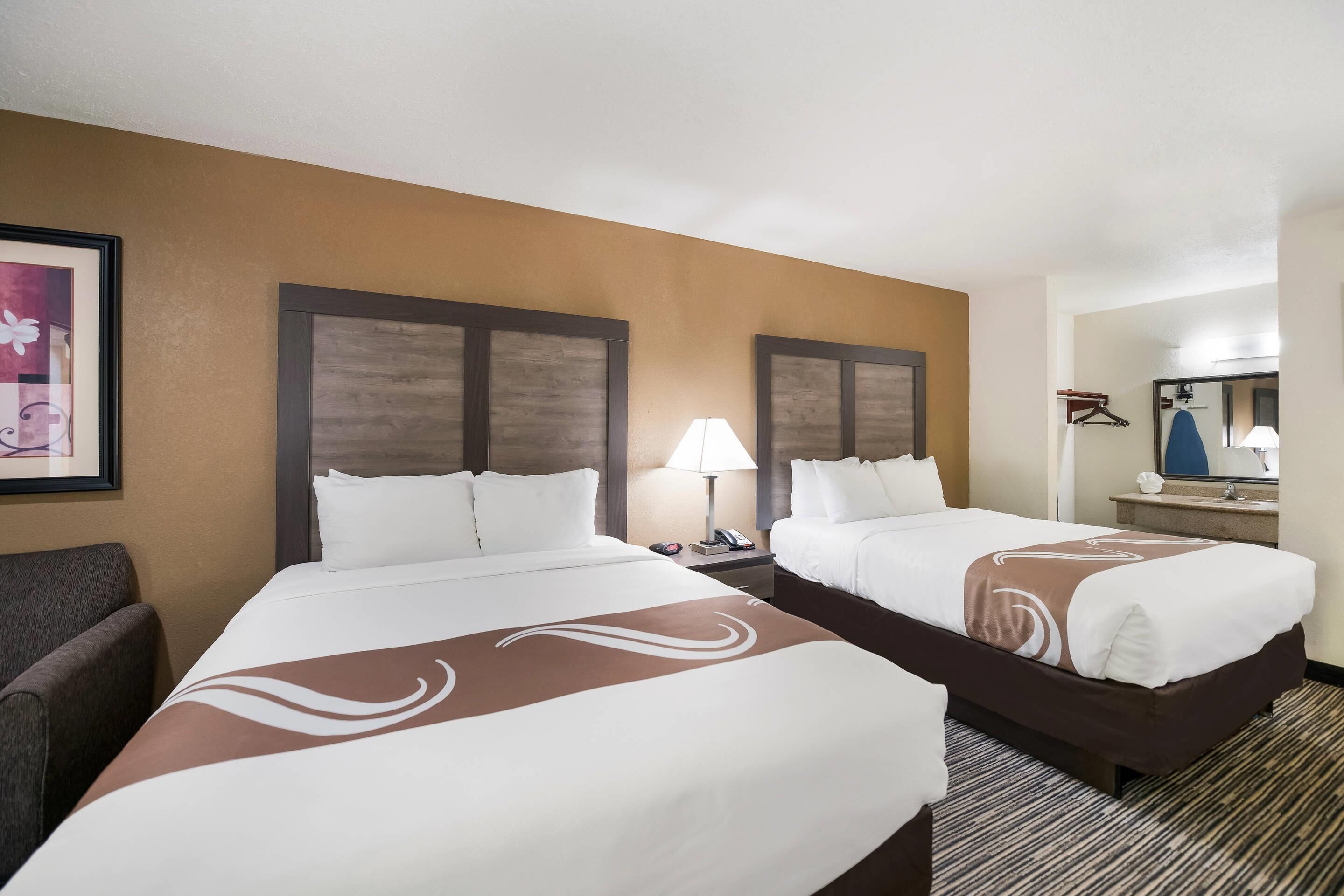 Une chambre d’hôtel avec deux lits, une table de chevet avec une lampe, une chaise, un miroir et un tableau encadré au mur.