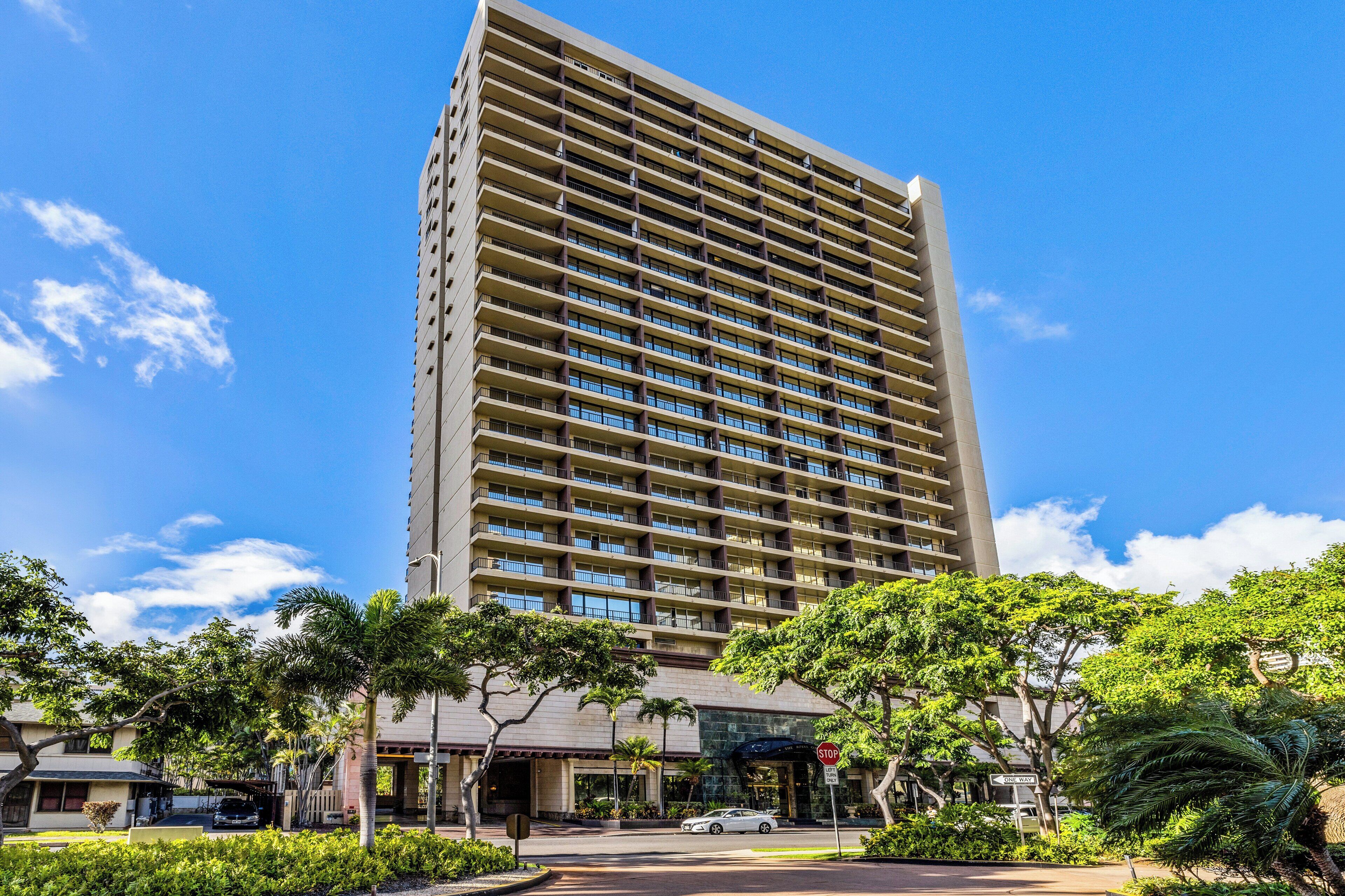 A tall hotel building with multiple floors and balconies, surrounded by greenery and a clear sky.
