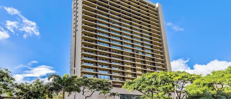 A tall hotel building with multiple floors and balconies, surrounded by greenery and a clear sky.