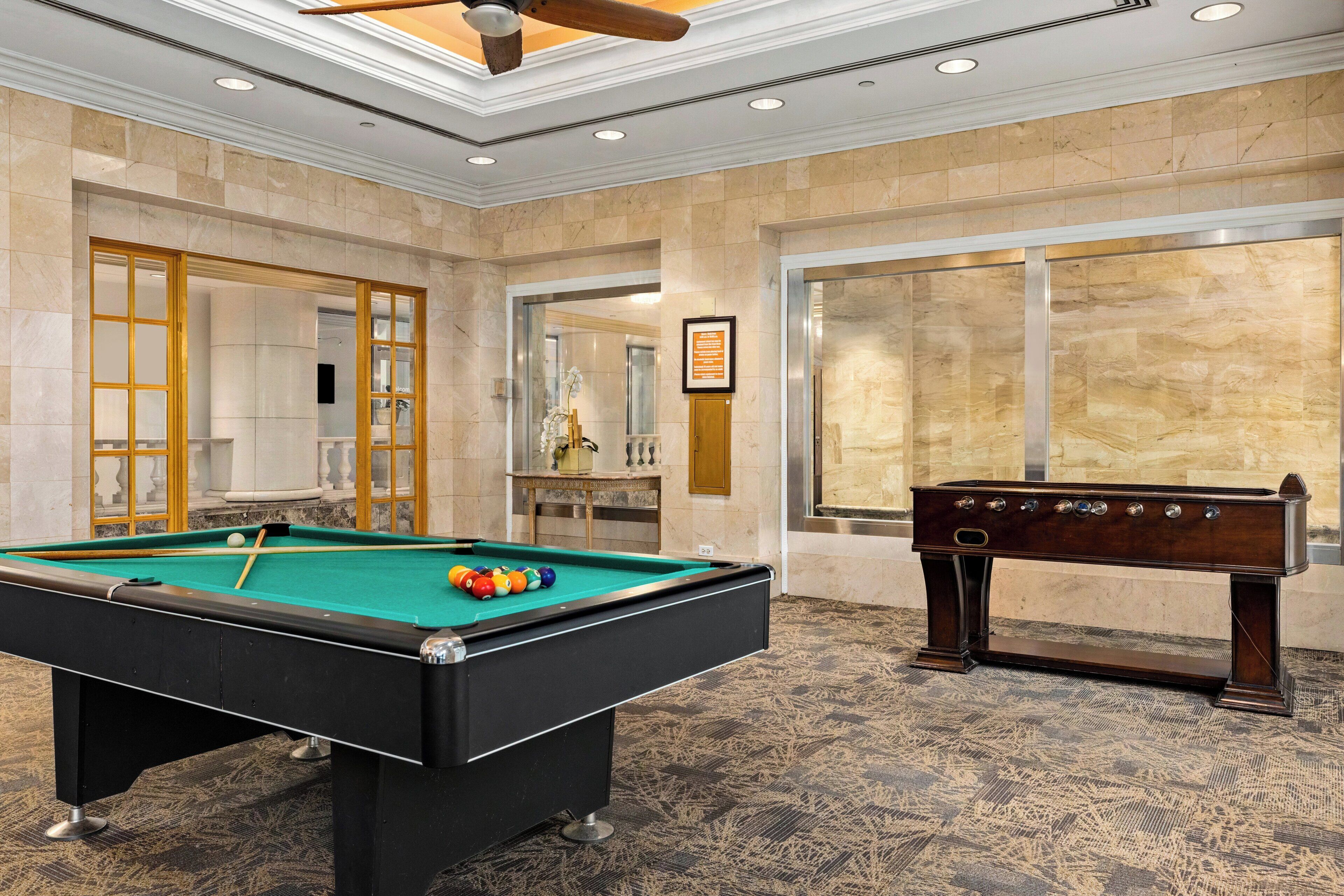 A pool table with balls and a foosball table in a room with marble walls and a ceiling fan.