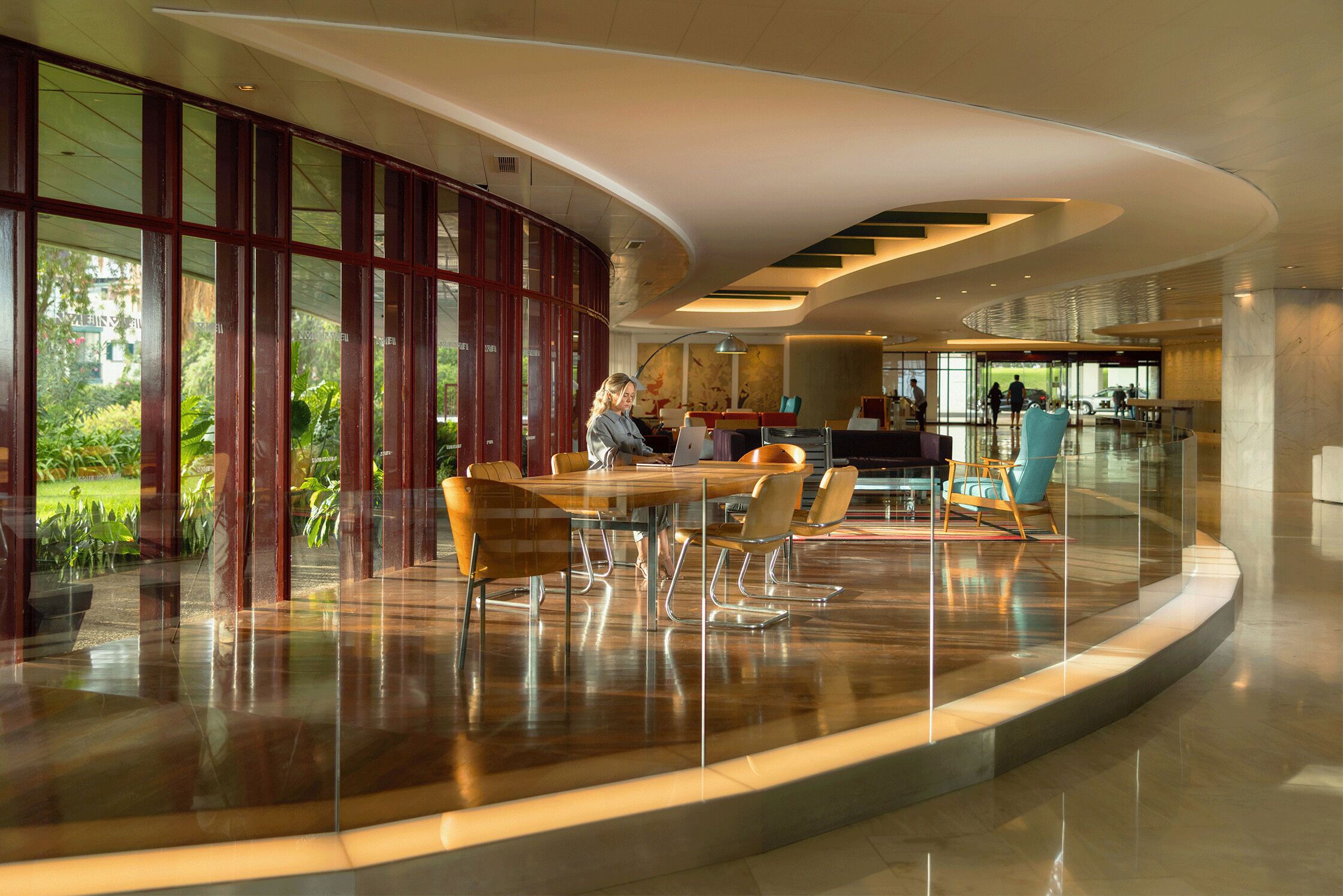 Eine moderne Hotelhalle mit einer gewölbten Glasscheibe, einem Holztisch für Konferenzen und einer Sitzecke mit Stühlen.