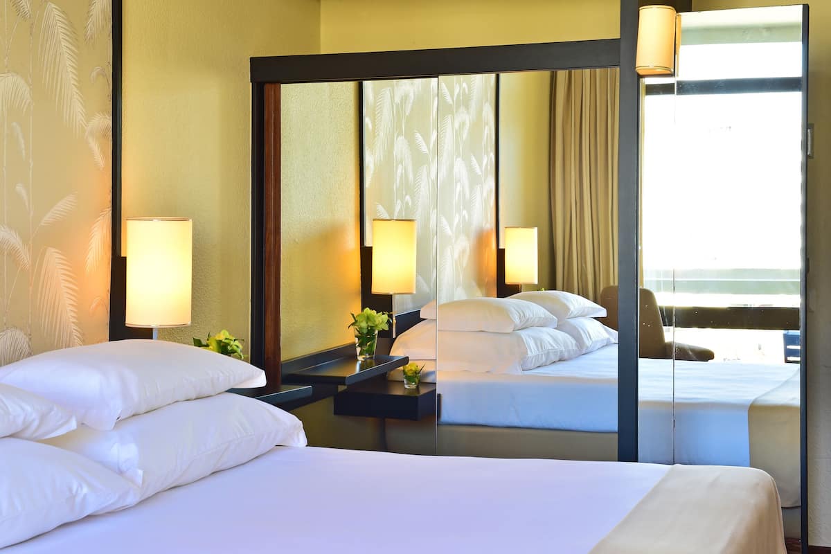 Une chambre d’hôtel avec un grand lit, une table de chevet avec une lampe, un miroir et une vue sur la mer.
