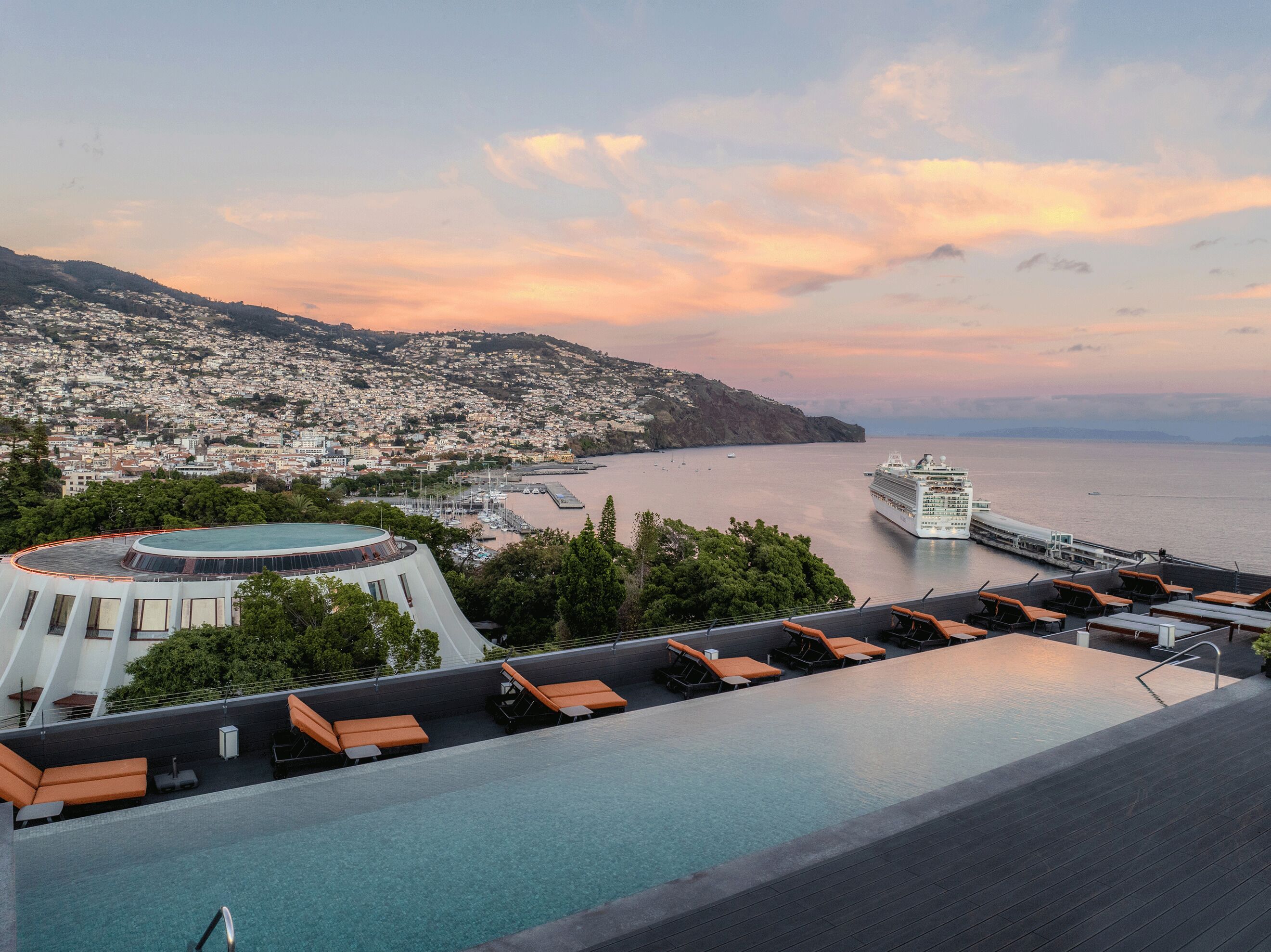 Una piscina infinita en la azotea con tumbonas, vista a un crucero y un paisaje urbano con colinas al fondo.