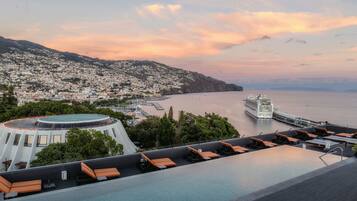 Una piscina infinita en la azotea con tumbonas, vista a un crucero y un paisaje urbano con colinas al fondo.