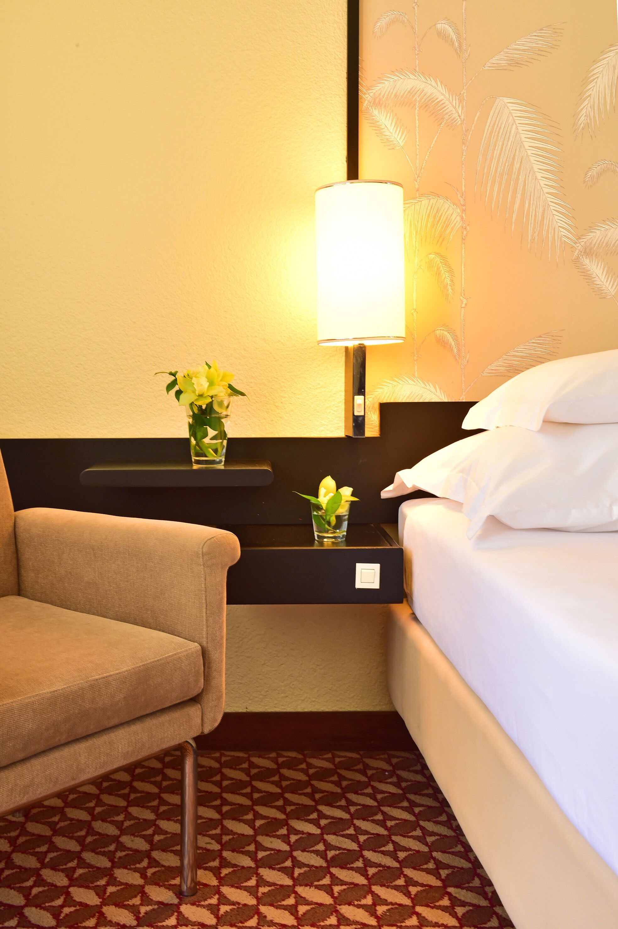 A hotel room with a bed, a chair, a nightstand with a lamp, and a wall with palm tree wallpaper.