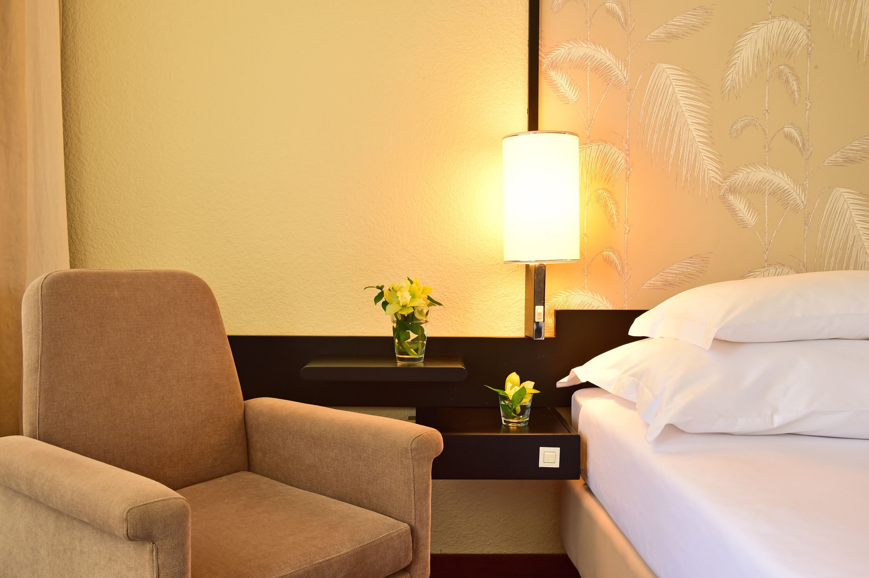 A hotel room with a bed, a chair, a nightstand with a lamp, and a wall with a palm leaf pattern.