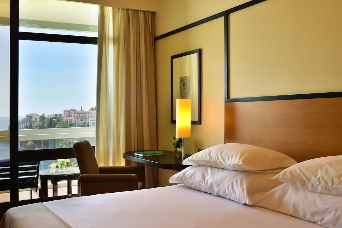 Une chambre d’hôtel avec un grand lit, un bureau avec une lampe, une chaise et une vue sur la ville grâce à de grandes fenêtres.