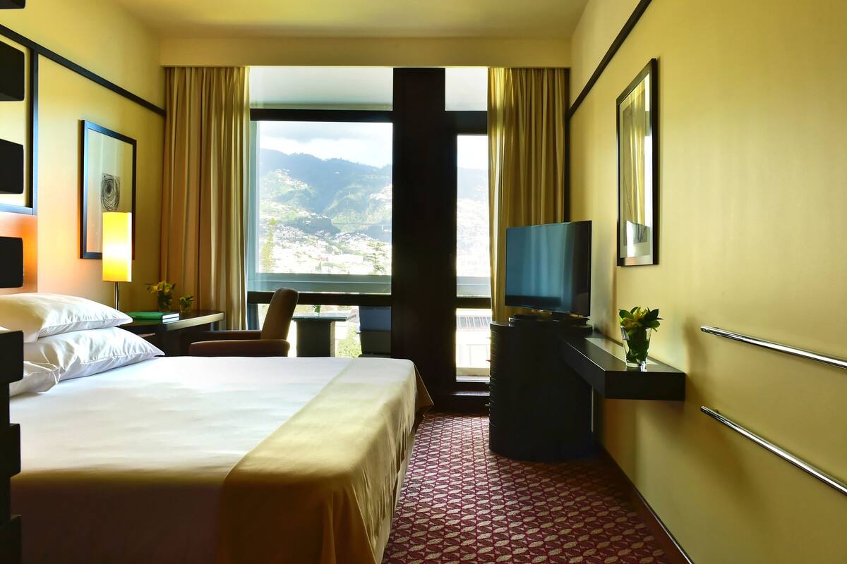 Une chambre d’hôtel comprenant un lit, un bureau avec une chaise, une télévision et une grande fenêtre donnant sur la montagne.