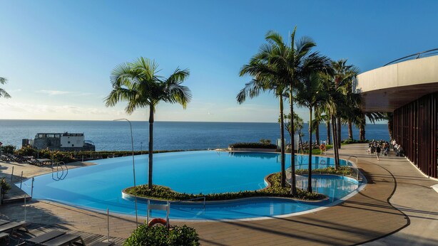 A pool with palm trees, a view of the ocean, and a building with a curved roof. - Pestana Casino Park Ocean and SPA Hotel (Funchal)