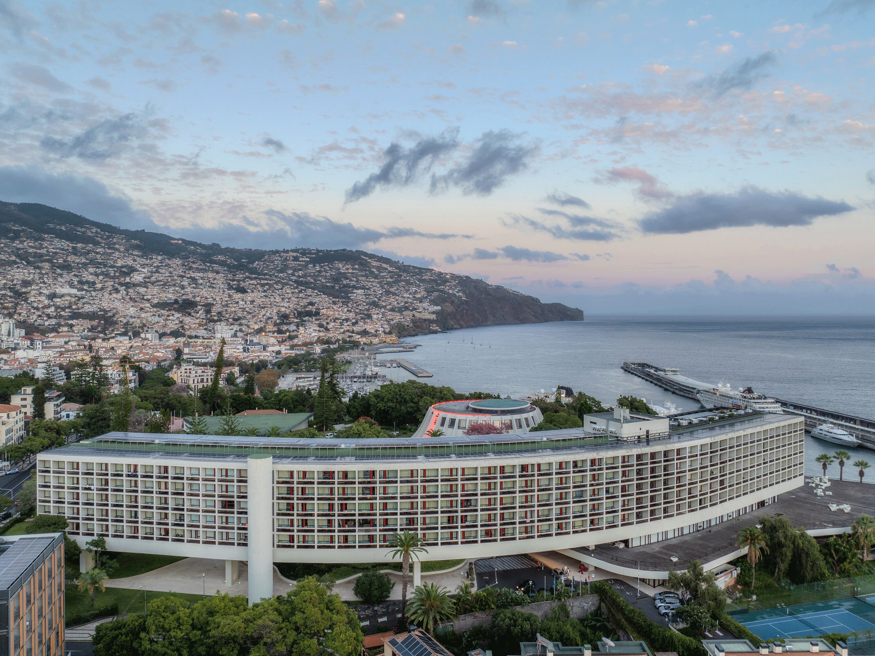 A modern hotel with a curved facade, located near a coastal cityscape with a view of the sea and a hilly background.