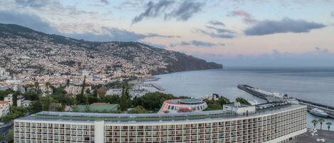 A modern hotel with a curved facade, located near a coastal cityscape with a view of the sea and a hilly background.