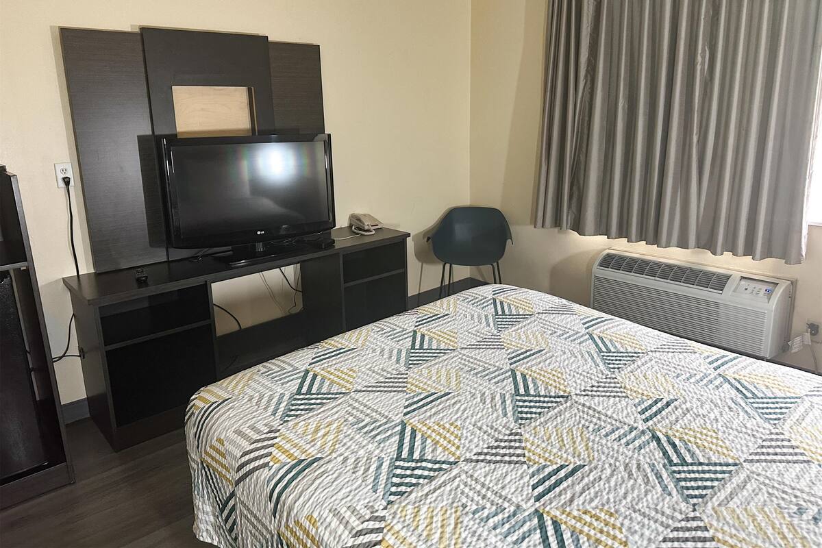 A hotel room with a bed, a TV on a stand, a chair, and an air conditioning unit.